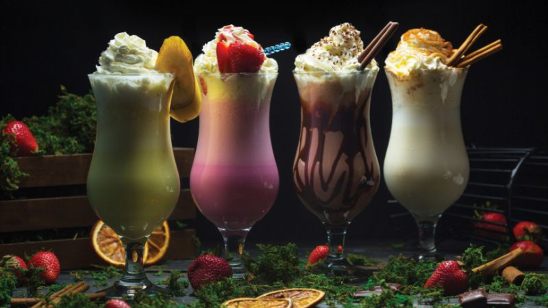 Four colorful milkshakes topped with whipped cream and fresh fruit garnishes in glass serving glasses