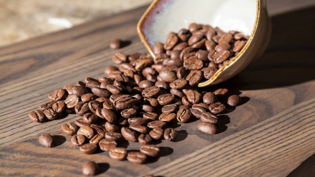 Roasted coffee beans scattered on a wooden table with a ceramic bowl in the background.
